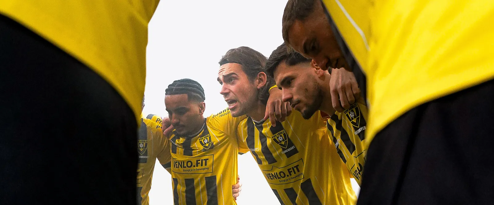 Spelers van VVV-Venlo in een huddle, gefotografeerd tussen twee geel-zwarte shirts door
